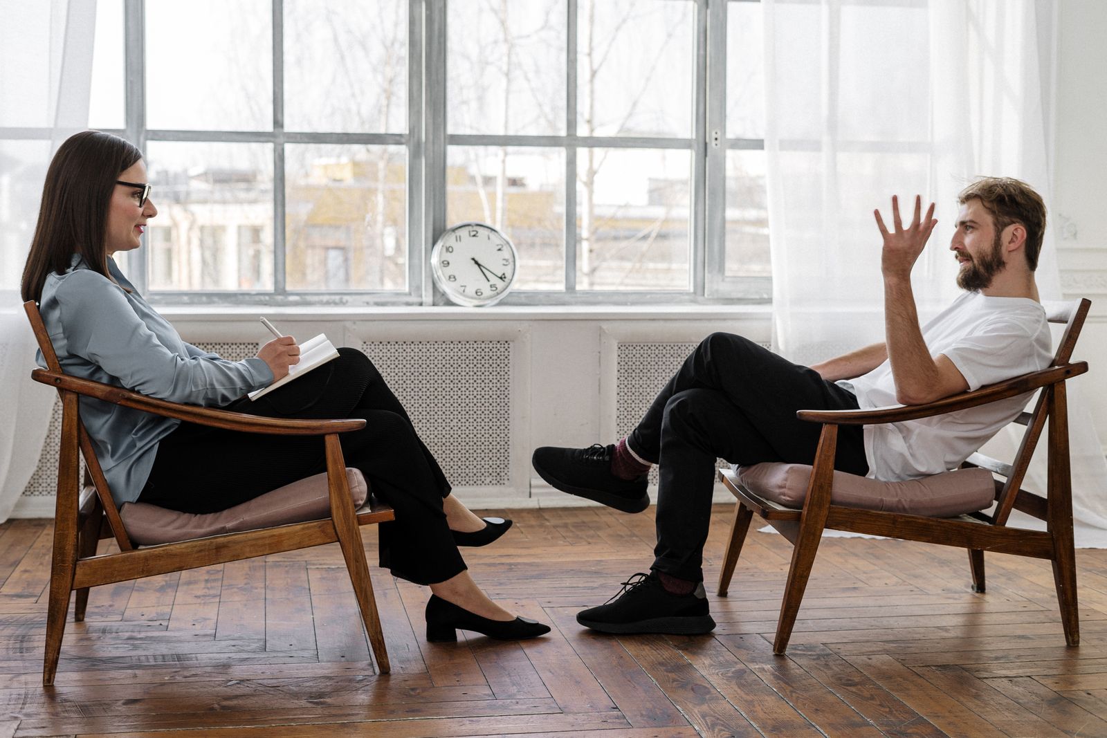 A man and a woman sit facing each other in a bright room, talking while she takes notes